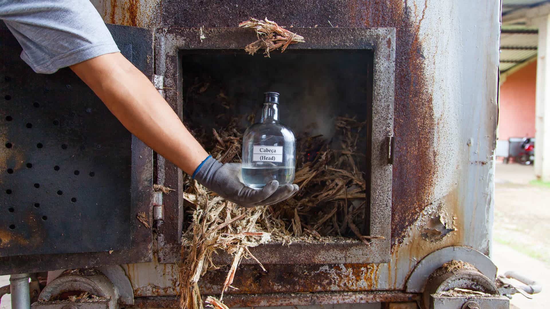 man holding bottle of cachaça head in front of a bagaço-burning furnace